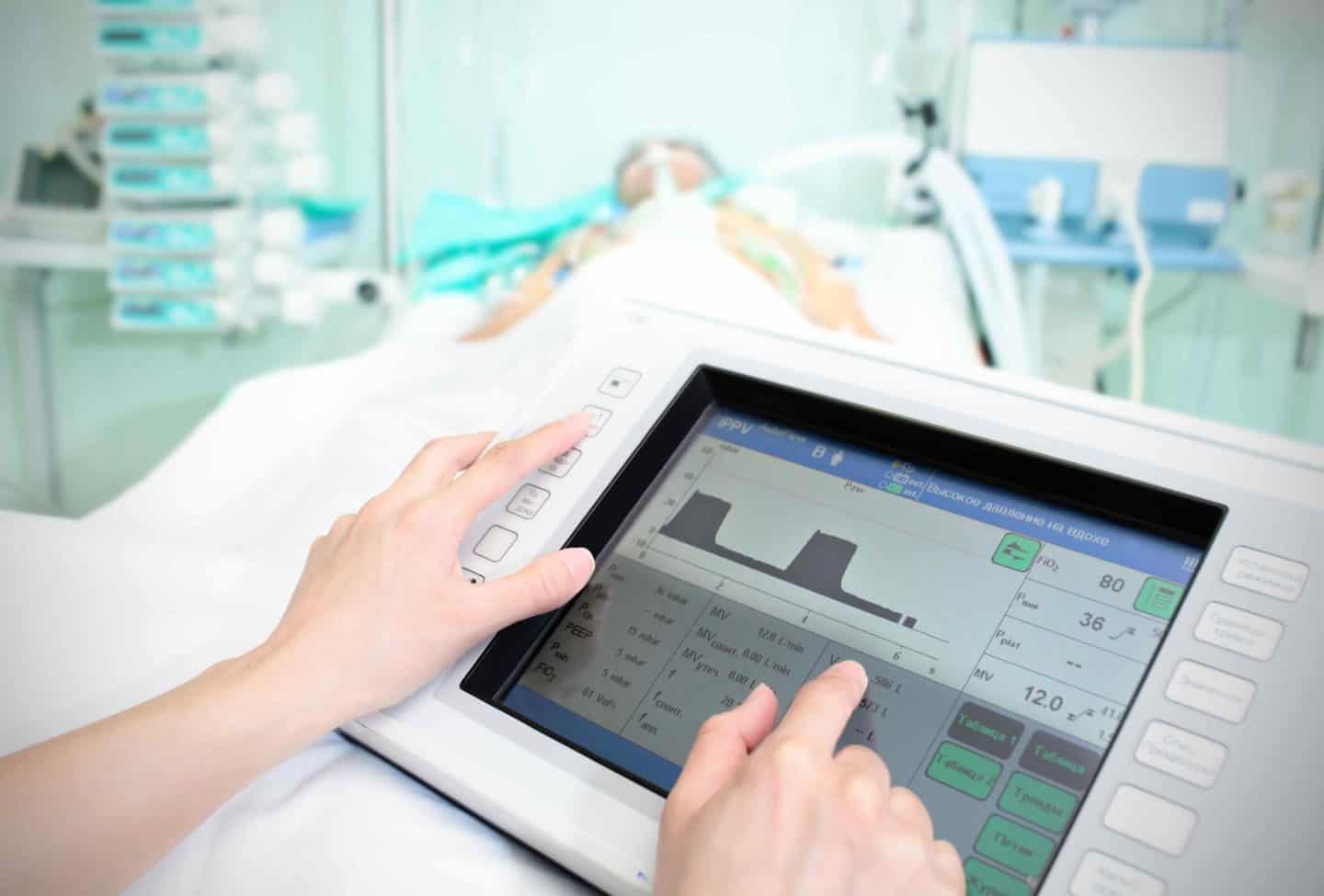 A hospital worker's hands using a monitoring device in front of an ICU patient on a ventilator